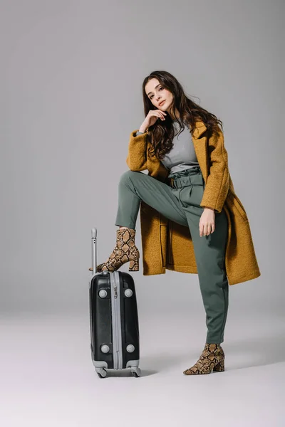 Donna alla moda in cappotto beige in posa con borsa da viaggio su grigio — Foto stock