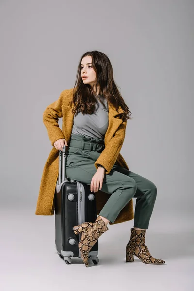 Fashionable girl in beige coat sitting on travel bag on grey — Stock Photo