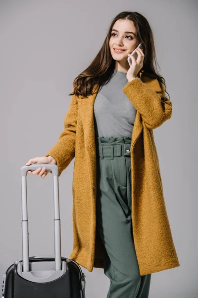 Smiling woman in beige coat posing with travel bag and talking on smartphone, isolated on grey — Stock Photo