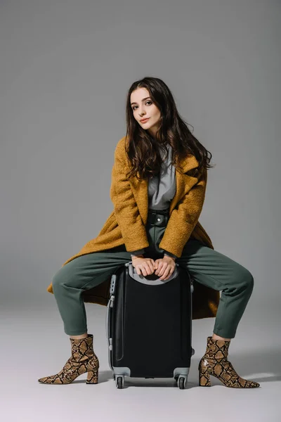 Attractive woman in beige coat sitting on travel bag on grey — Stock Photo
