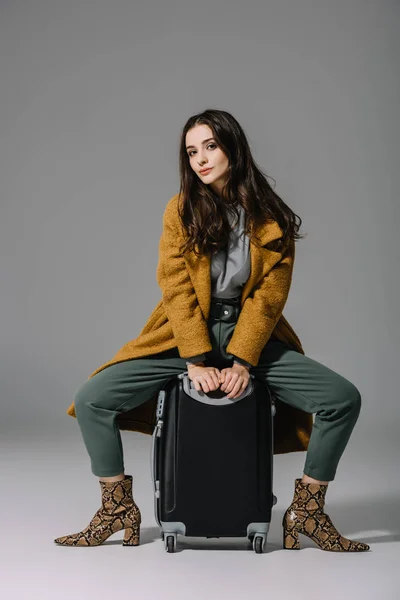 Attraente donna in cappotto beige seduta su borsa da viaggio su grigio — Foto stock