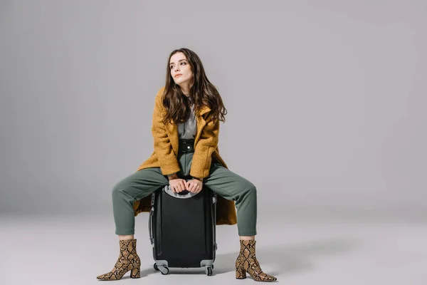 Elegante ragazza alla moda in cappotto beige seduta su borsa da viaggio su grigio — Foto stock