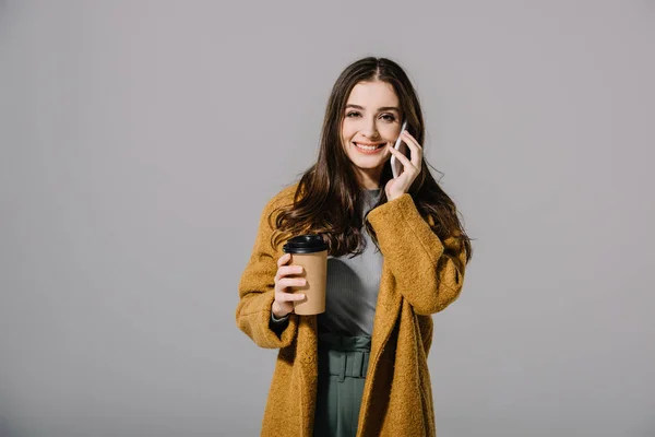 Симпатичная улыбчивая девушка в бежевом пальто, держащая в руках кофе, пока разговаривает по смартфону, изолированная на сером — стоковое фото