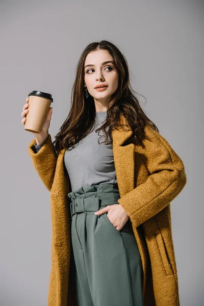 Attractive girl in beige coat holding coffee to go, isolated on grey — Stock Photo