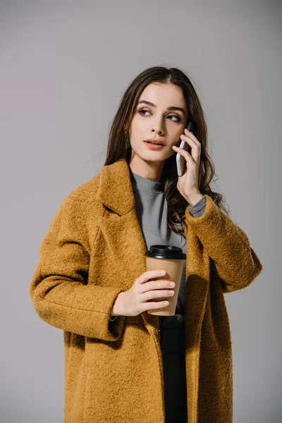 Menina bonita no casaco bege segurando café para ir enquanto fala no smartphone, isolado no cinza — Fotografia de Stock