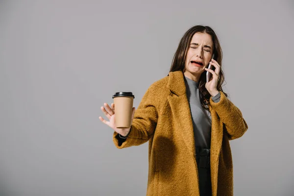 Triste llorando chica en beige abrigo sosteniendo café para ir mientras habla en el teléfono inteligente, aislado en gris - foto de stock
