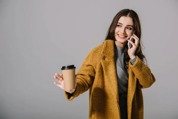 Lächelndes Mädchen im beigen Mantel, Kaffee to go haltend, während es auf dem Smartphone spricht, isoliert auf grau — Stockfoto