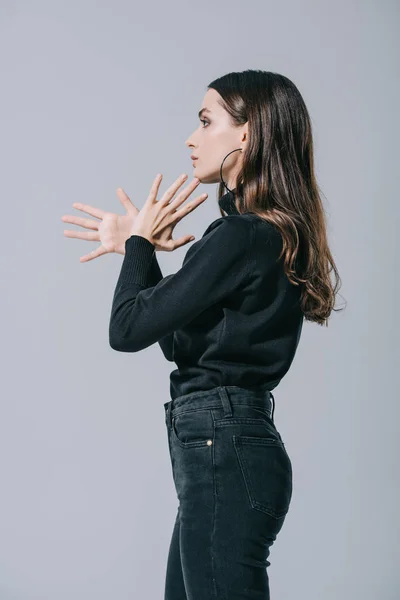 Vista laterale della ragazza alla moda gesticolando e posando in maglione nero, isolato su grigio — Foto stock