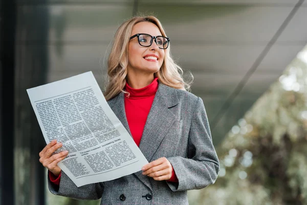 Attrayant femme d'affaires en manteau et lunettes souriant et tenant journal — Photo de stock