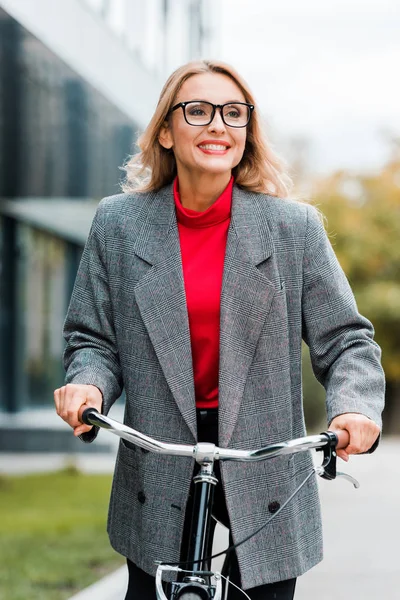 Attractive businesswoman in coat and glasses smiling and riding bike — Stock Photo