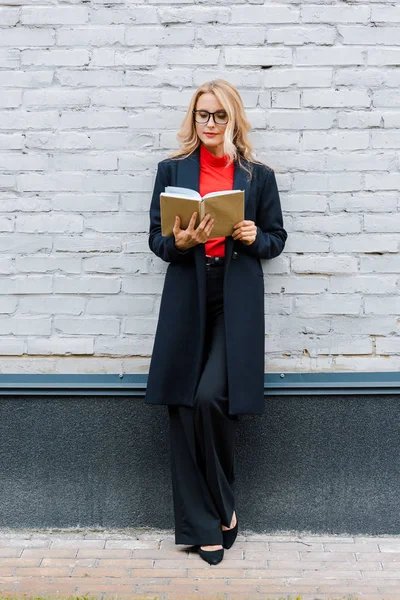 Atractiva mujer de negocios en abrigo negro y gafas libro de lectura - foto de stock