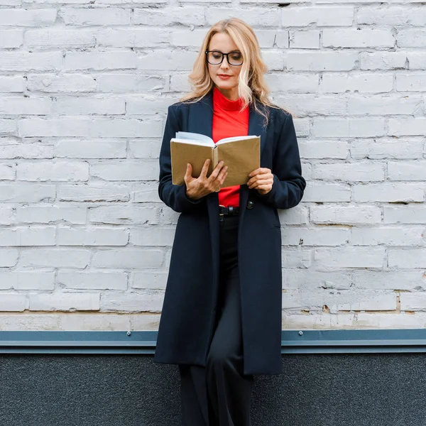 Attraktive Geschäftsfrau in schwarzem Mantel und Brille liest Buch — Stockfoto