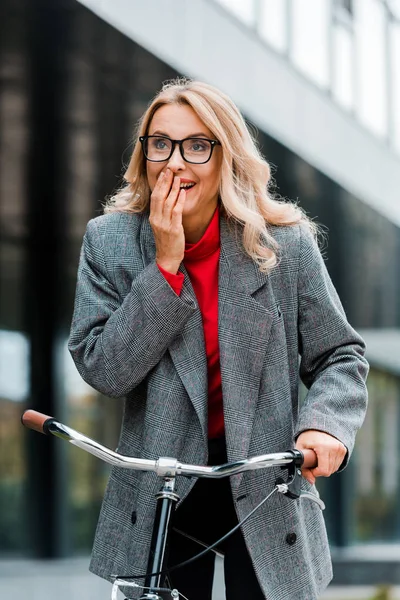 Atractiva mujer de negocios en abrigo y gafas sonriendo y montar en bicicleta - foto de stock