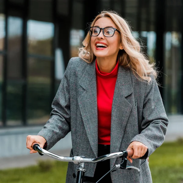 Atractiva mujer de negocios en abrigo y gafas sonriendo y montar en bicicleta - foto de stock