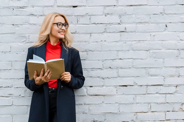 Attraktive Geschäftsfrau in Mantel und Brille, lächelnd und mit Buch in der Hand — Stockfoto