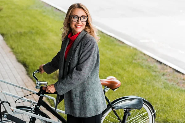 Attractive businesswoman in coat and glasses smiling and holding bike — Stock Photo