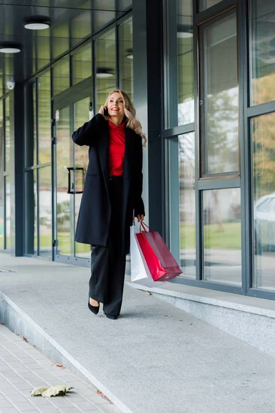 Atractiva mujer de negocios hablando en el teléfono inteligente y sosteniendo bolsas de compras - foto de stock