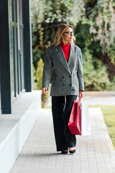 Atractiva mujer de negocios en abrigo y gafas caminando y sosteniendo bolsas de compras - foto de stock
