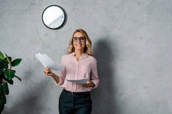 Mulher de negócios atraente em camisa e óculos sorrindo e segurando papéis — Fotografia de Stock