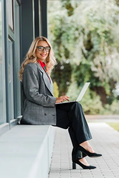 Séduisante femme d'affaires en manteau et lunettes souriant et tenant ordinateur portable — Photo de stock