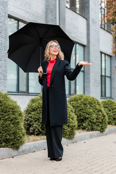 Atractiva mujer de negocios en abrigo y gafas con paraguas negro con la mano extendida - foto de stock