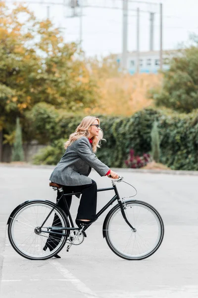 Side view of attractive businesswoman in coat and glasses riding bike — Stock Photo