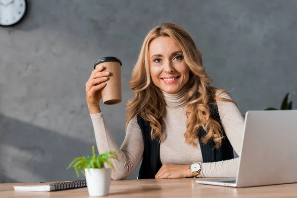 Приваблива бізнес-леді посміхається і тримає паперовий стаканчик в офісі — стокове фото