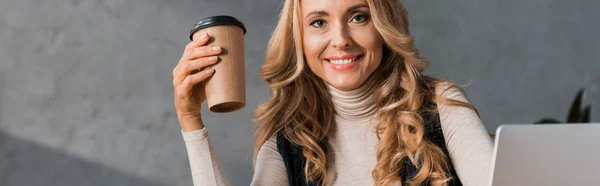 Panoramic shot of attractive businesswoman smiling and holding paper cup in office — Stock Photo