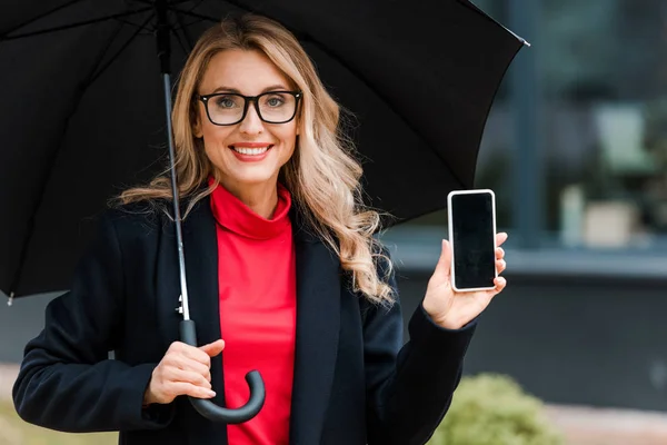 Femme d'affaires attrayante et souriante en manteau tenant parapluie et smartphone — Photo de stock