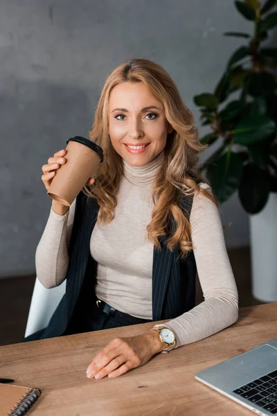 Atractiva y rubia empresaria sentada en la mesa y sosteniendo la taza de papel - foto de stock