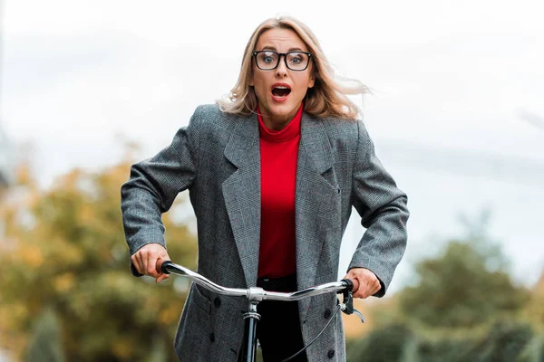 Attractive and shocked businesswoman in grey coat riding bike — Stock Photo
