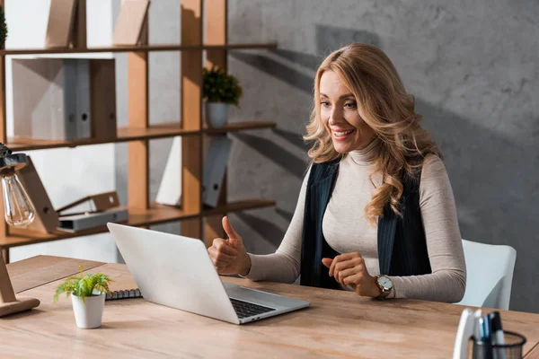 Atraente e sorridente empresária mostrando polegar para cima e olhando para laptop — Fotografia de Stock