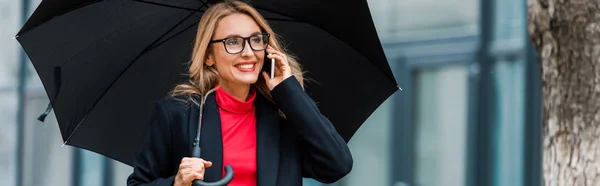 Panoramaaufnahme einer attraktiven und lächelnden Geschäftsfrau in schwarzem Mantel, die einen Regenschirm hält und mit dem Smartphone spricht — Stockfoto