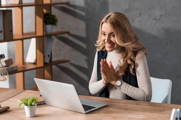 Atraente e sorridente empresária mostrando as mãos orando e olhando para laptop — Fotografia de Stock