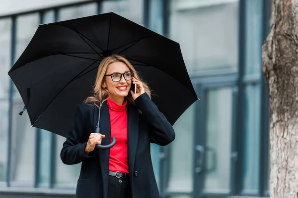 Atraente e sorridente empresária em casaco preto segurando guarda-chuva e falando no smartphone — Fotografia de Stock