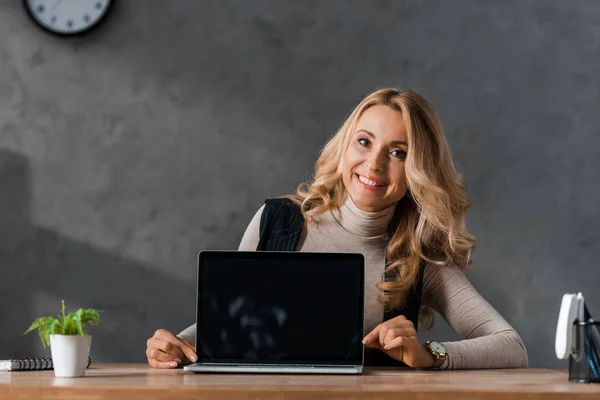 Attraente e sorridente donna d'affari seduta a tavola e con in mano un computer portatile con schermo bianco — Foto stock