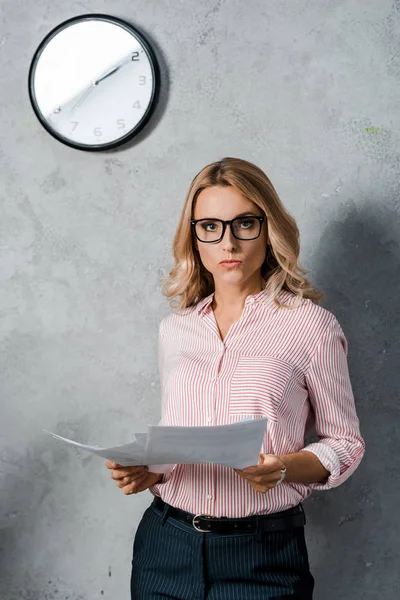Attractive businesswoman in glasses and shirt holding papers in office — Stock Photo