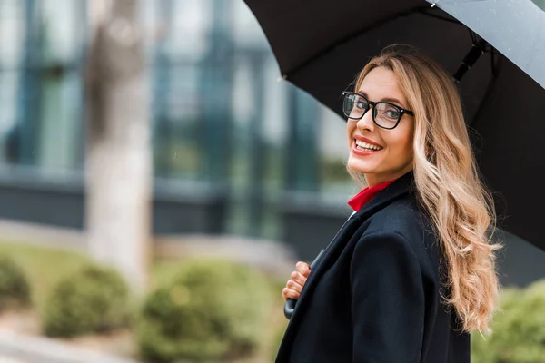 Attractive and smiling businesswoman in black coat holding umbrella — Stock Photo