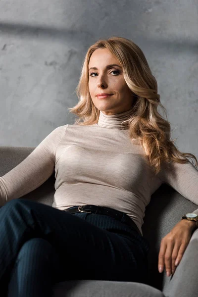 Attractive and blonde businesswoman sitting on armchair and looking at camera — Stock Photo