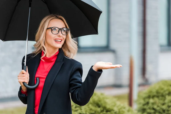 Attraente e sorridente donna d'affari in cappotto nero con mano tesa che tiene ombrello — Foto stock