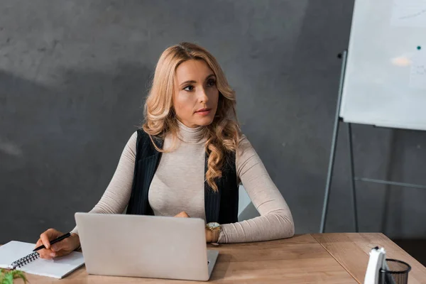 Attraktive und blonde Geschäftsfrau, die am Tisch sitzt und wegschaut — Stockfoto