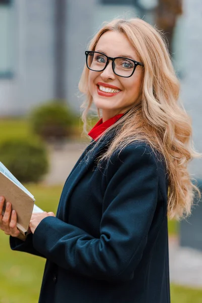 Attraktive und lächelnde Geschäftsfrau in schwarzem Mantel und Brille mit Buch — Stockfoto