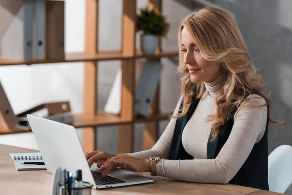 Femme d'affaires attrayante et blonde assise à table et utilisant un ordinateur portable — Photo de stock