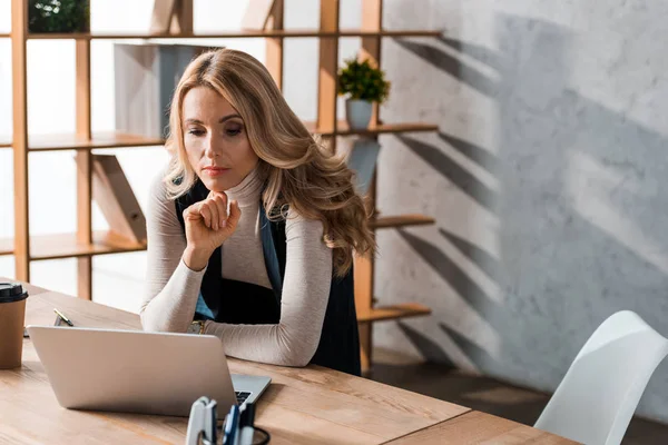 Attractive and blonde businesswoman sitting at table and looking at laptop — Stock Photo