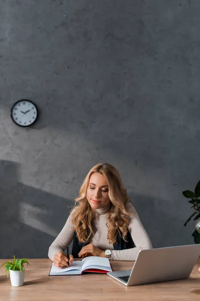Приваблива і блондинка бізнес-леді сидить за столом і пише в блокноті — стокове фото