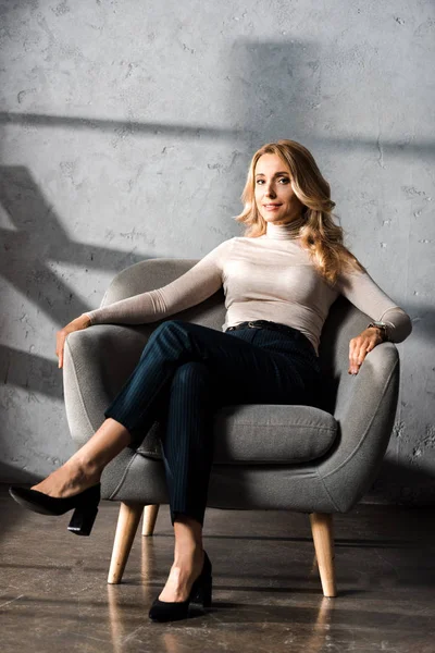 Attractive and blonde businesswoman smiling and sitting on armchair in office — Stock Photo