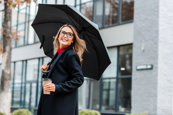 Attractive and blonde woman in black coat holding paper cup and umbrella — Stock Photo