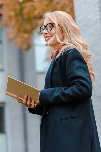 Attractive and blonde businesswoman in black coat smiling and holding book — Stock Photo