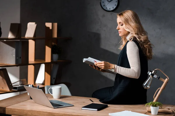 Attractive and blonde businesswoman holding papers and sitting on table in office — Stock Photo