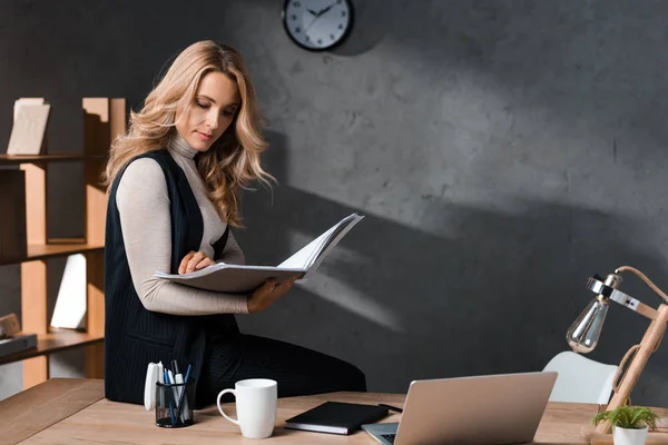 Attraktive und blonde Geschäftsfrau, die Papiere hält und im Büro auf dem Tisch sitzt — Stockfoto